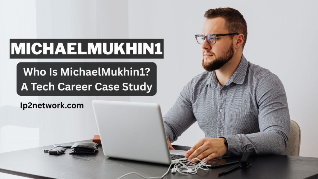 A man with short hair, a beard, and glasses sits at a desk working on a laptop. He is wearing a long-sleeve button-up shirt and looking to the side. On the desk are a smartphone, a wallet, keys, and some cables. Text on the image reads “MICHAELMUKHIN1 – Who Is MichaelMukhin1? A Tech Career Case Study – lp2network.com.”