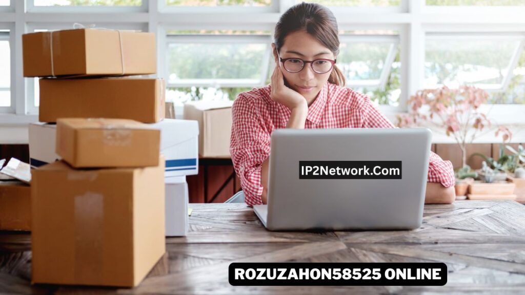 A woman wearing glasses and a red checkered shirt sits at a wooden table, looking at a laptop with a focused expression. Several cardboard shipping boxes are stacked around her, suggesting an online business or packaging workspace. Text on the laptop reads “IP2Network.com,” and a bold label at the bottom says “ROZUZAHON58525 ONLINE.”